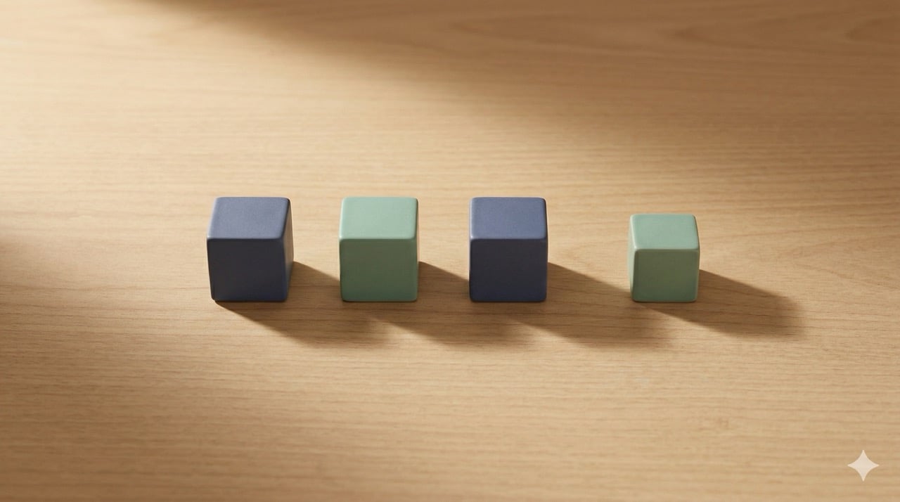 Four small geometric cubes in slate blue and sage green arranged in a deliberate row on a warm wood surface, casting soft shadows, representing a curated, intentional selection. 
